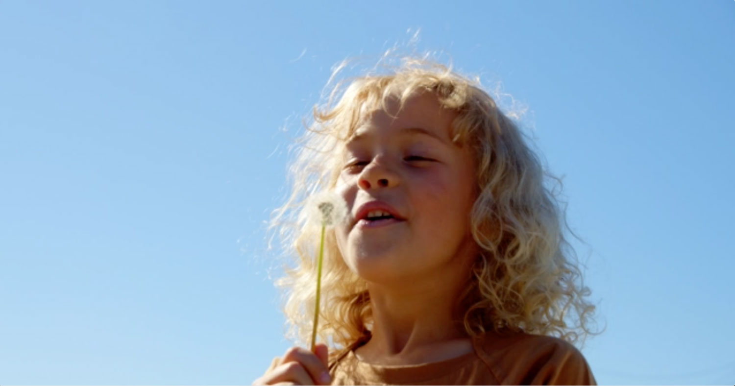 A reconnected child with curly blonde hair, blowing a dandelion under a bright blue sky