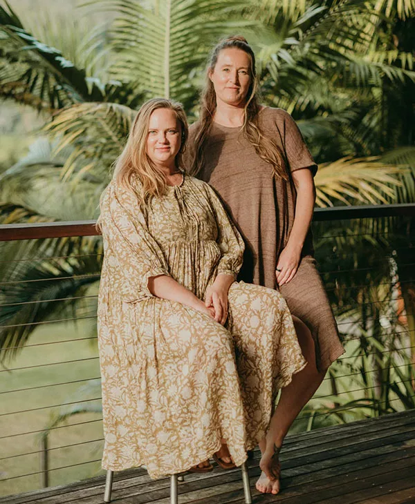 Emma & Eleanor side-by-side on a timber deck, surrounded by palm trees, smiling gently